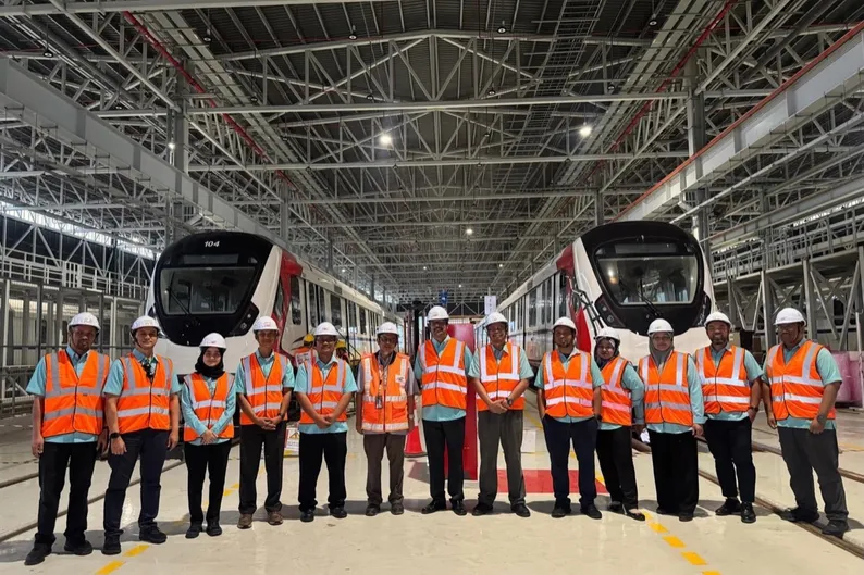 CIDB Malaysia and CREAM delegates in high-visibility vests and hard hats standing between two RTS Link trains at Wadi Hana Depot