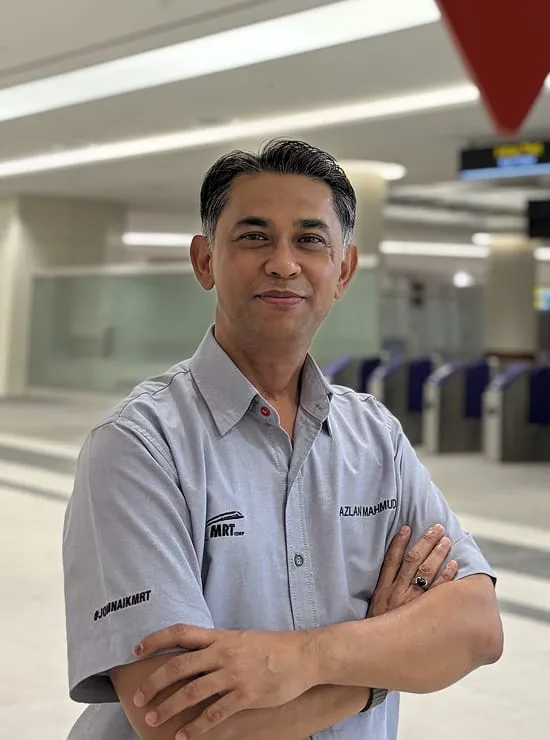 A man wearing MRT corporate attire standing in the Bukit Chagar station concourse, with the ticket gates behind him