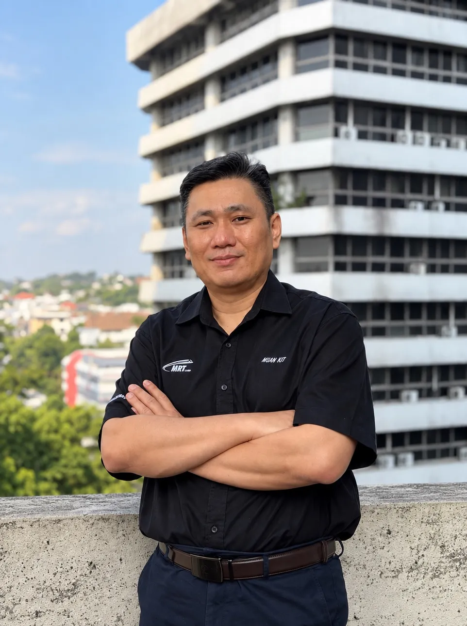 Tan Mun Kit in a black MRT Corp shirt, arms crossed, standing on a rooftop overlooking the city skyline