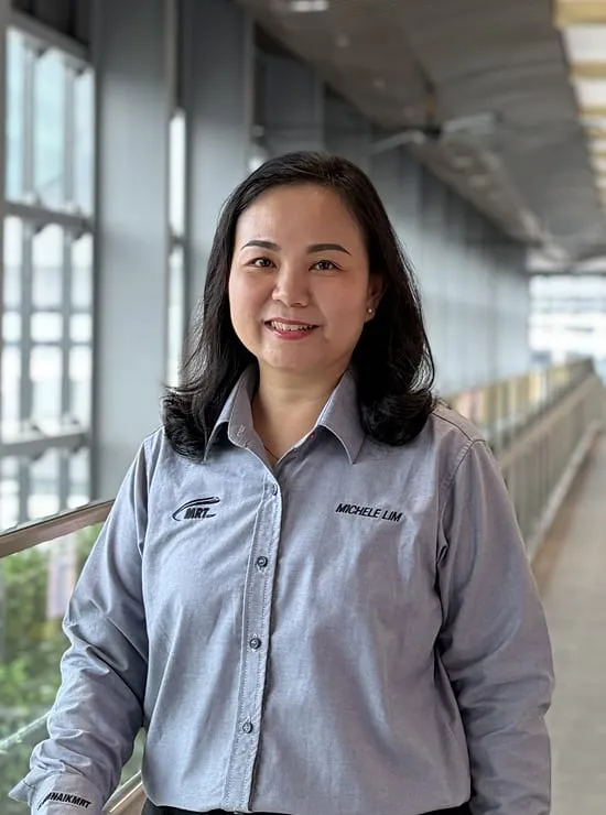 A woman wearing MRT corporate attire standing in a hallway, with a glass wall looking out to the outdoors behind her