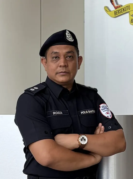 A man in auxiliary police uniform standing in front of a booth, with the Malaysian coat of arms on the wall behind him