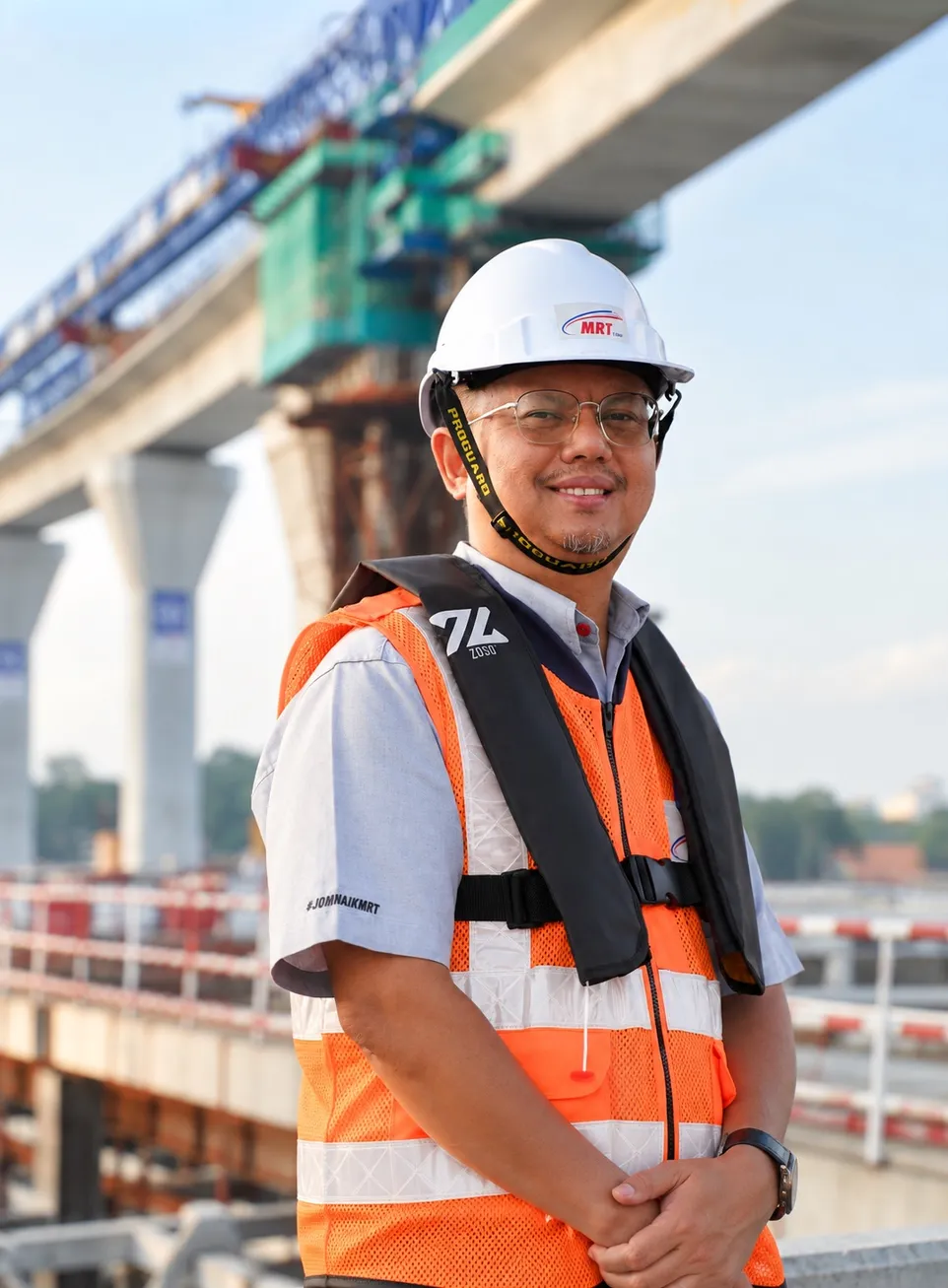 Ir. Zulkifli Mohamed in a white MRT Corp hard hat and orange high-visibility vest, smiling at a viaduct construction site with a crane in the background